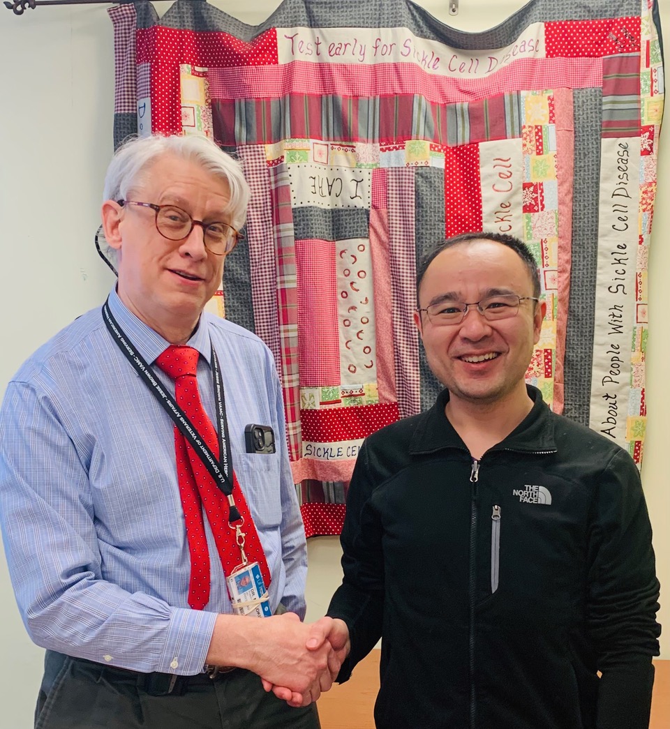 Robert Molokie of Division of Hematology and Oncology at UI Health and Richard and Loan Hill Department of Biomedical Engineering Assistant Professor and member of the Center for Bioinformatics and Quantitative Biology Zhangli Peng shake hands in front of a quilt made by a sickle cell patient.